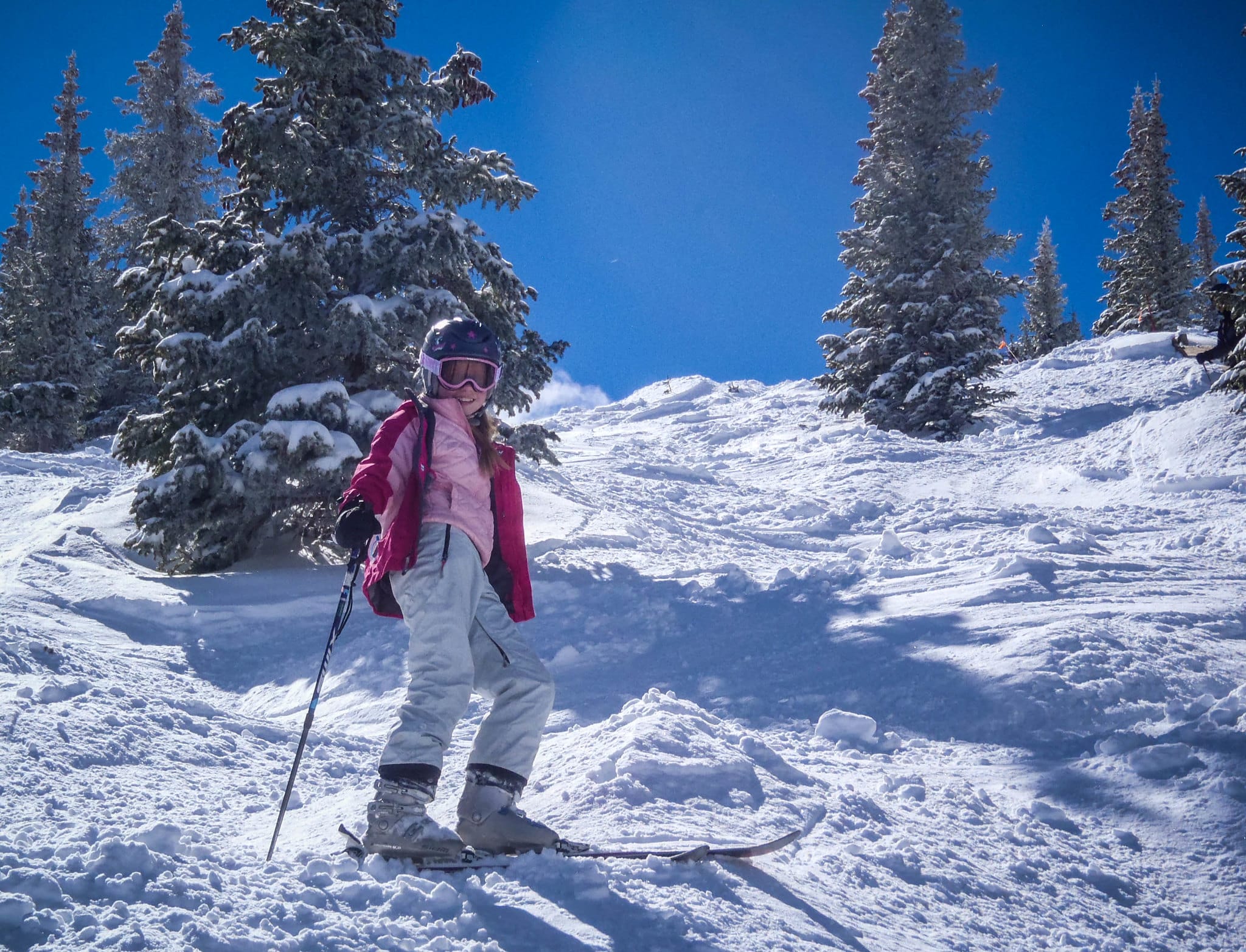Person in a pink ski jacket and white pants standing on the side of a snowy Breckenridge hill on skis, smiling