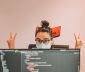 Woman in black rimmed glassed doing victory signs behind a computer display showing code