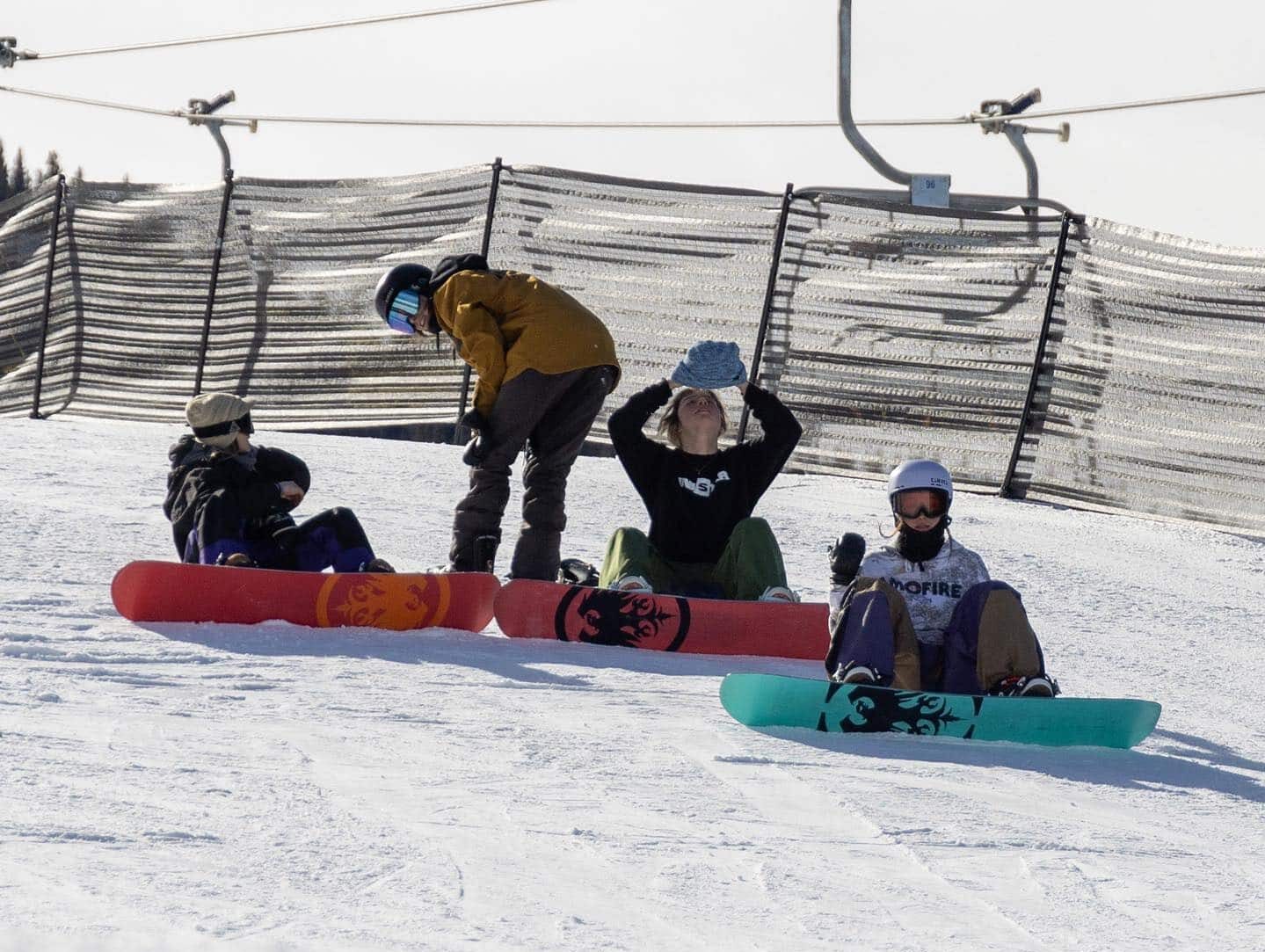 4 snowboarders sitting with their Never Summer board facing the camera at the bottom of a ski slope